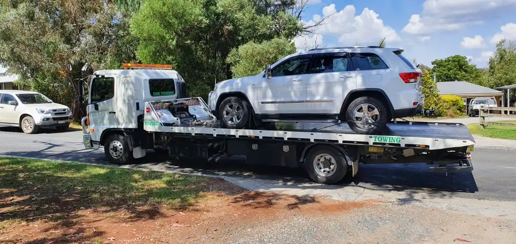 A white SUV loaded on a tilty tray tow truck parked on a suburban street, surrounded by trees and houses in the background.