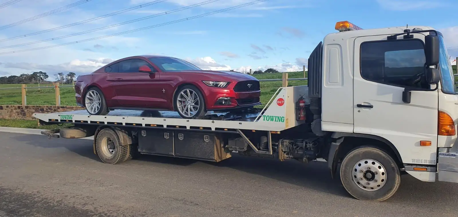A red Ford Mustang sports car being transported on a white tilty tray tow truck during the day, with a green field and blue sky in the background.