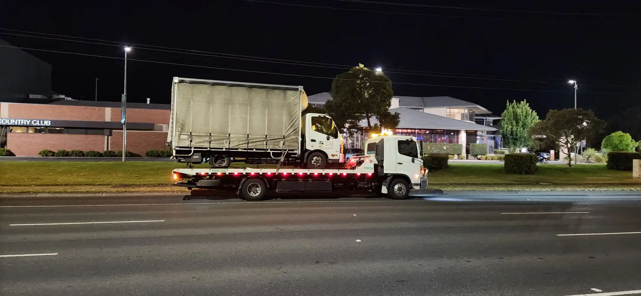 A white tilty tray tow truck carrying a large box truck at night, with a country club building visible in the background.