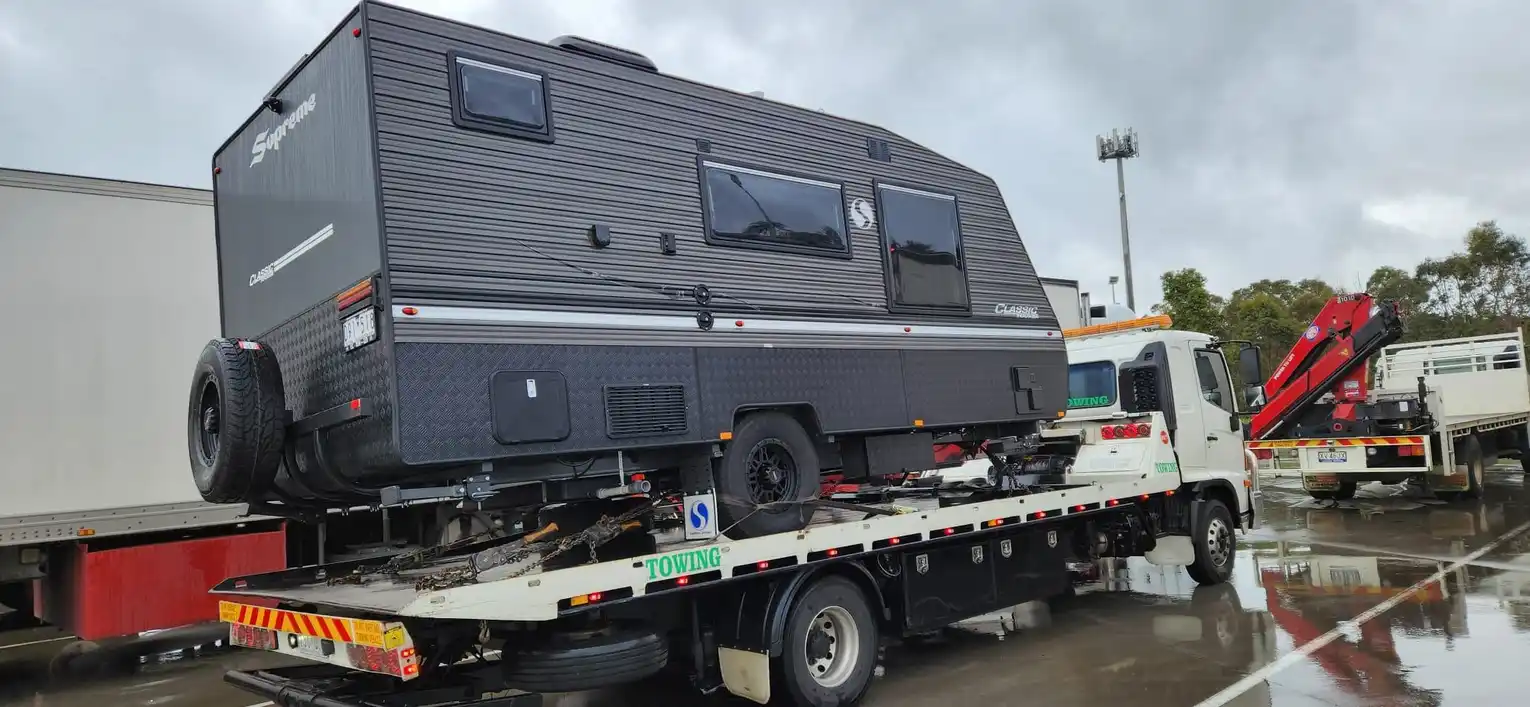 A white flatbed tow truck transporting a large black caravan in a wet, reflective parking lot, with other trucks and overcast skies in the background.
