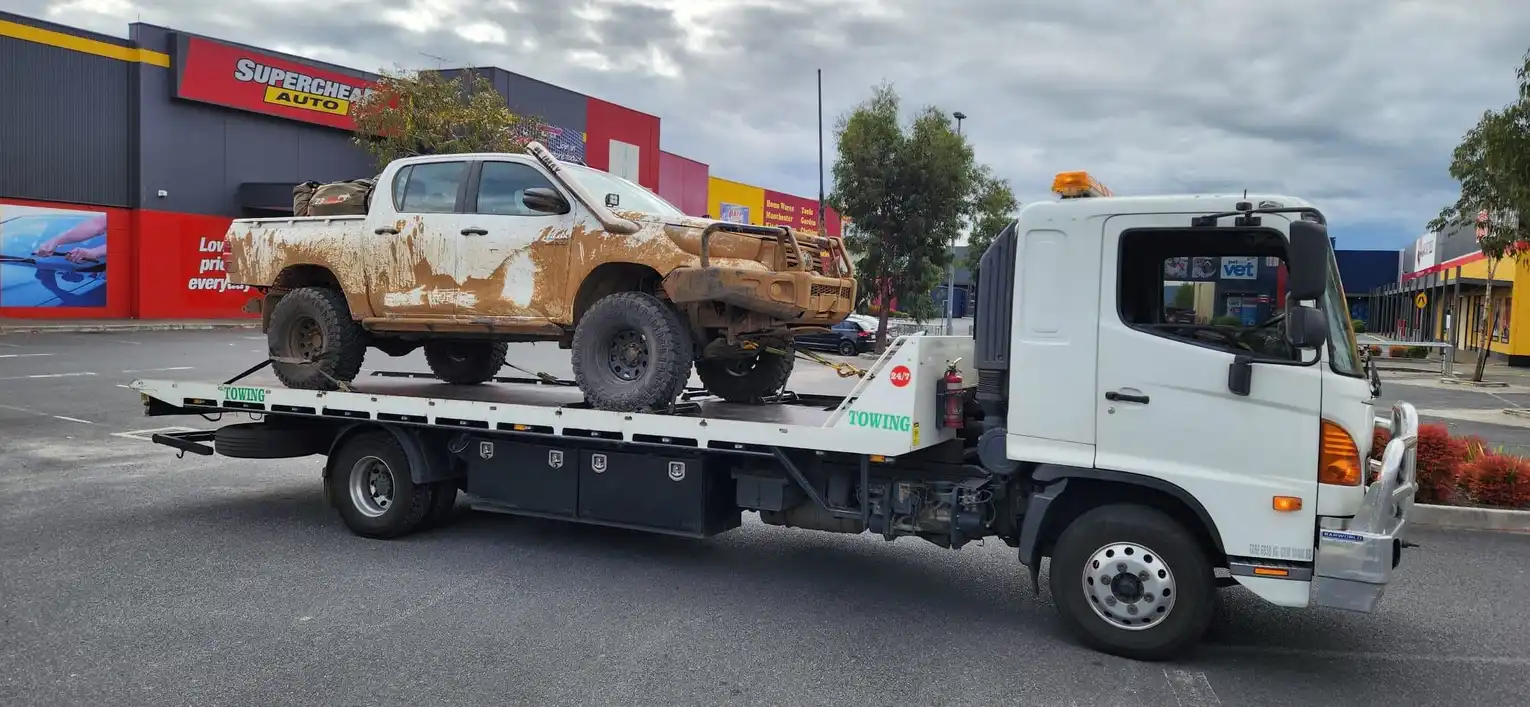 A white tilty tray tow truck transporting a mud-covered off-road utility vehicle, parked in front of an auto parts store in a commercial district.
