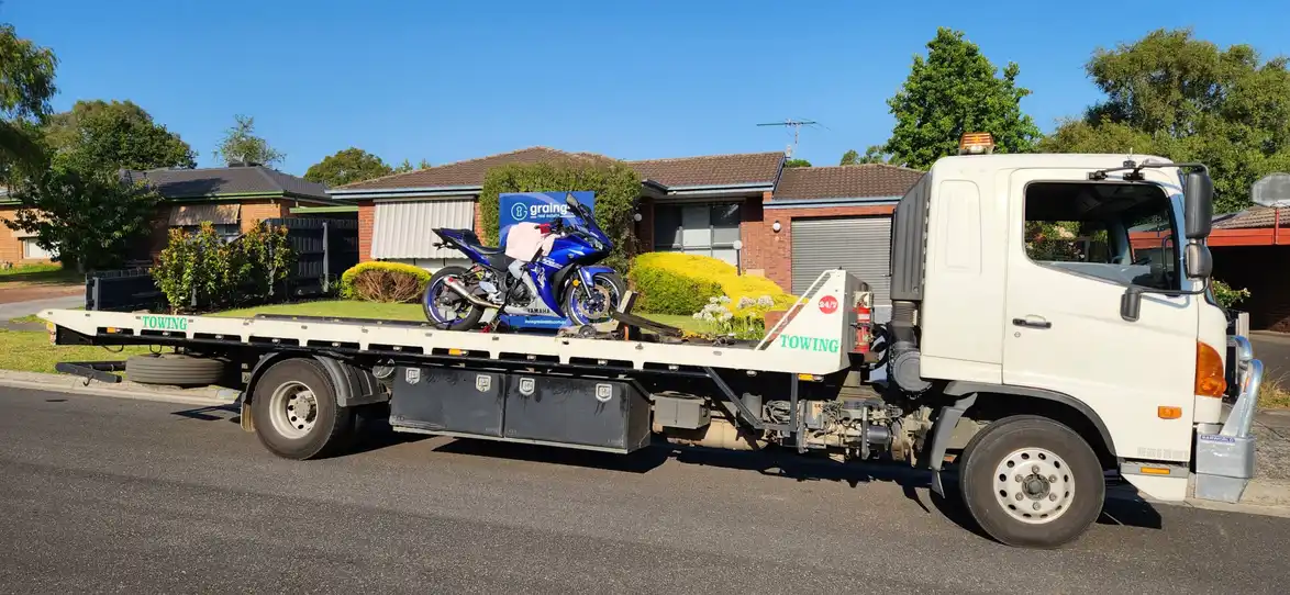 A white tilty tray tow truck carrying a blue Yamaha sports bike, parked on a suburban street in front of houses and gardens.