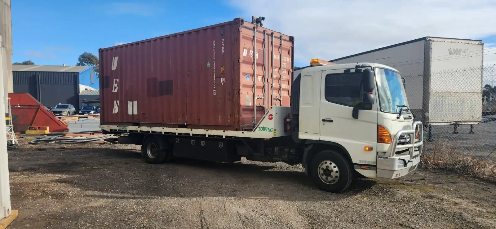 A white flatbed tow truck carrying a red shipping container, parked in a dirt lot surrounded by industrial buildings and equipment.