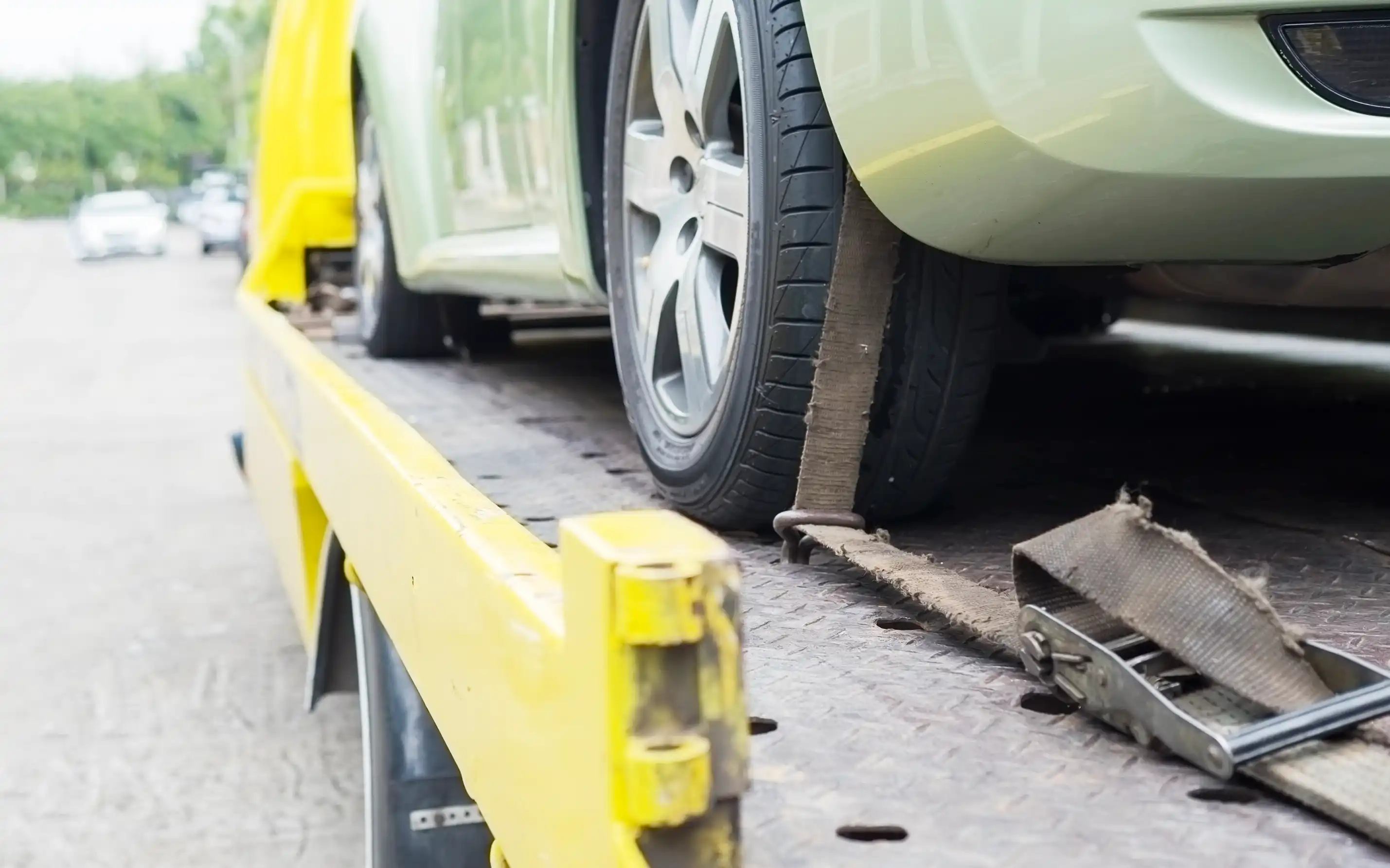A close-up of a car tyre secured with heavy-duty straps and ratchet buckles on a yellow flatbed transport truck showing proper load restraint.