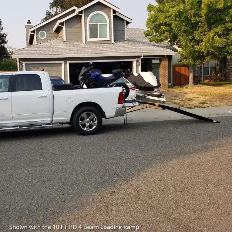 Motorcycle loaded into the bed of a pickup truck using a portable loading ramp, parked outside a suburban house.