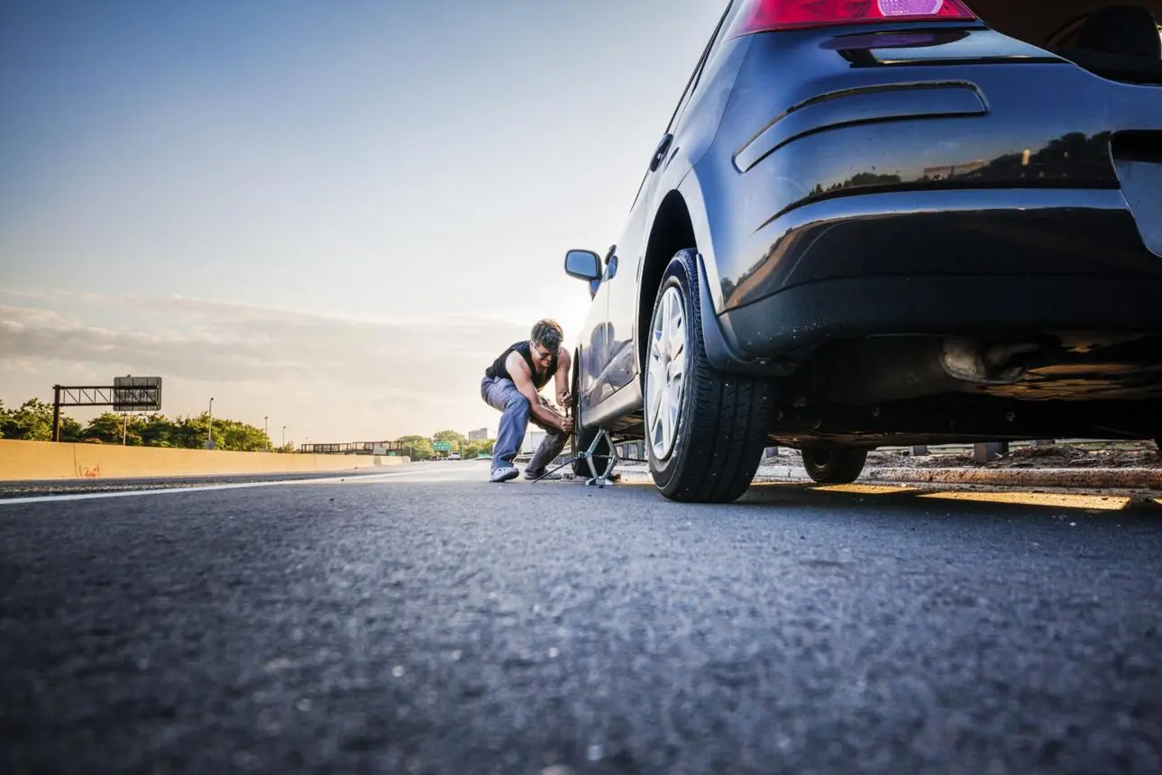 Driver standing beside a vehicle with a flat tyre on a busy highway shoulder while fast-moving traffic passes, showing the dangers of changing a tyre on the roadside.
