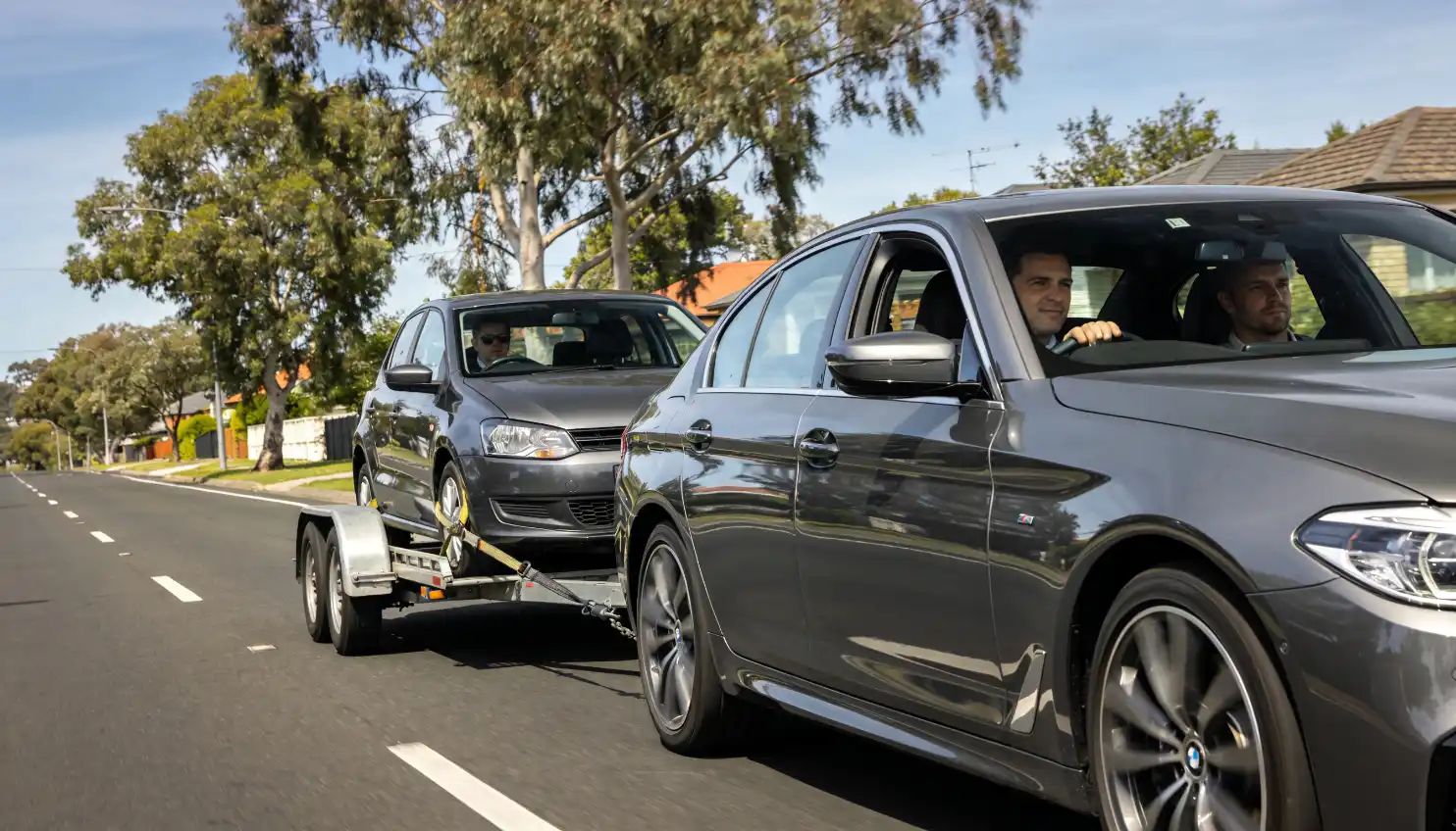 car towing another car on a trailer down a suburban street with two people visible in the towing vehicle